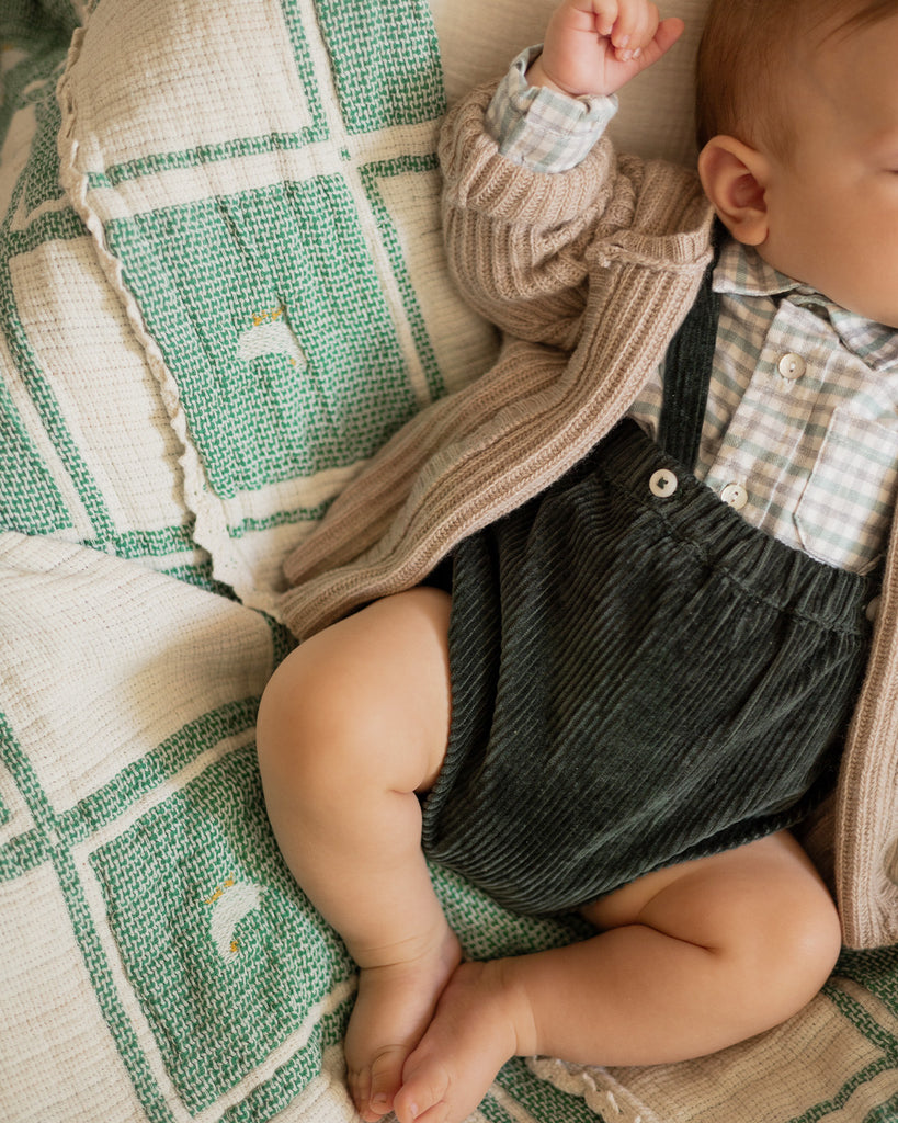 Bébé qui dort portant un bloomer vert sapin en velours associé à une chemise à carreaux et un gilet crème noisette