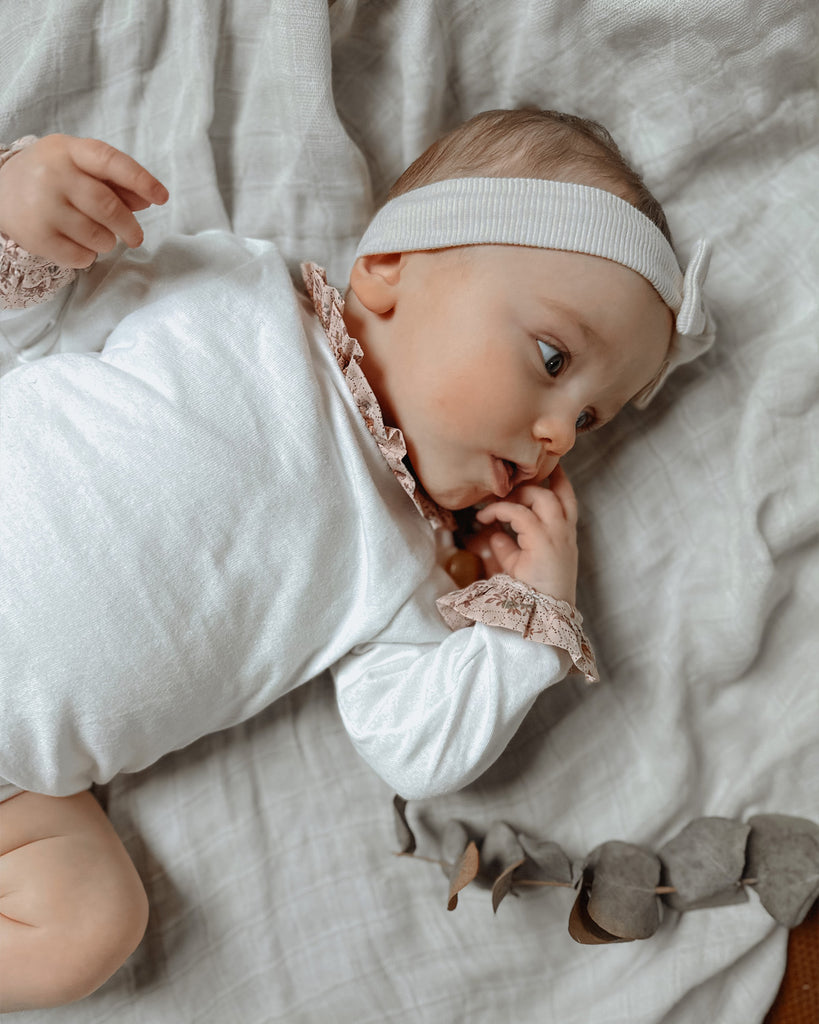 Bébé allongé sur un lit portant un bandeau à noeud de papillon blanc et un body manches longues à col et poignets volantés fleuris.