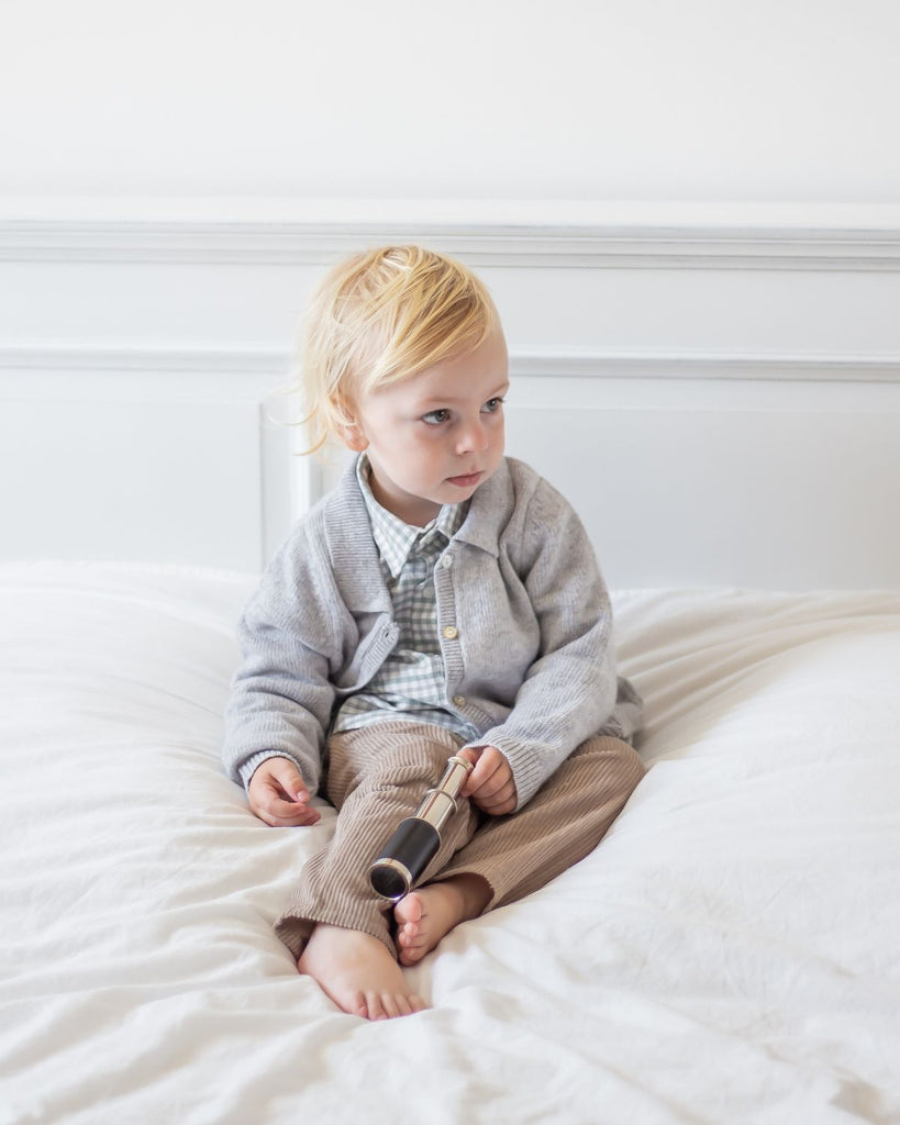 Petit garçon assis sur un lit portant un pantalon bebe en velours côtelé sable, une chemise à carreaux et un cardigan gris.