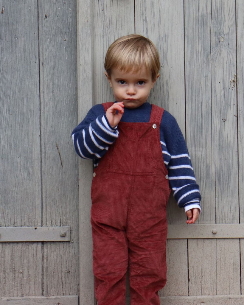 Petit garçon qui porte une salopette rouge en velours et un pull bleu à rayures blanches.