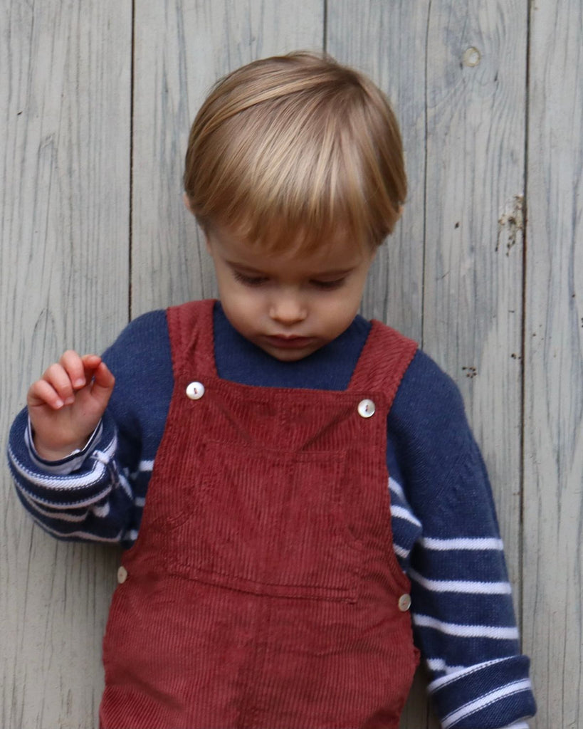 Petit garçon qui porte une salopette rouge en velours et un pull bleu à rayures blanches.