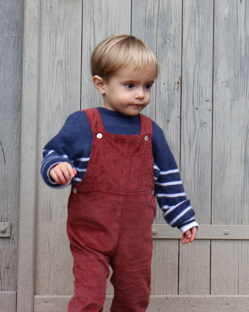 Petit garçon qui porte une salopette rouge en velours et un pull bleu à rayures blanches.