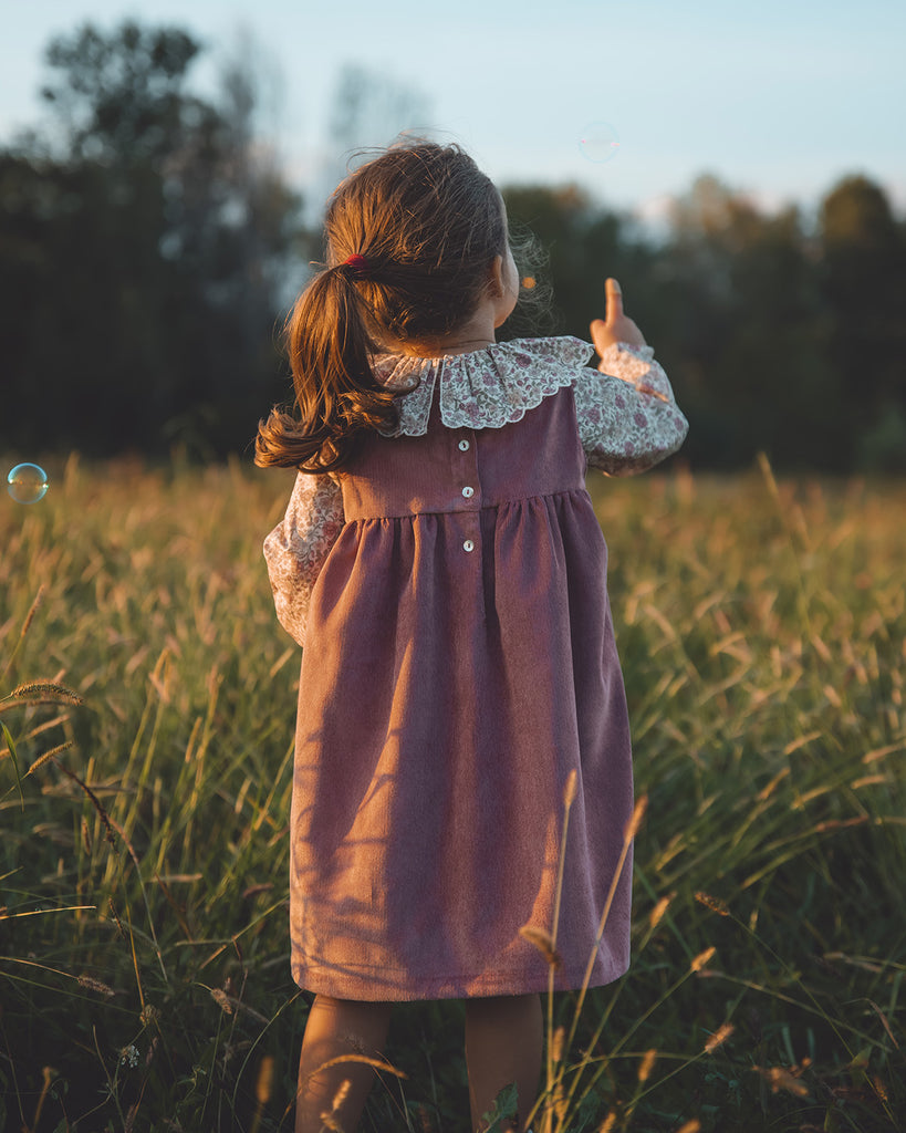 Vue de dos d'une petite fille debout dans un pré vêtue d'une robe en velours mauve avec une chemise fleurie à col volanté.