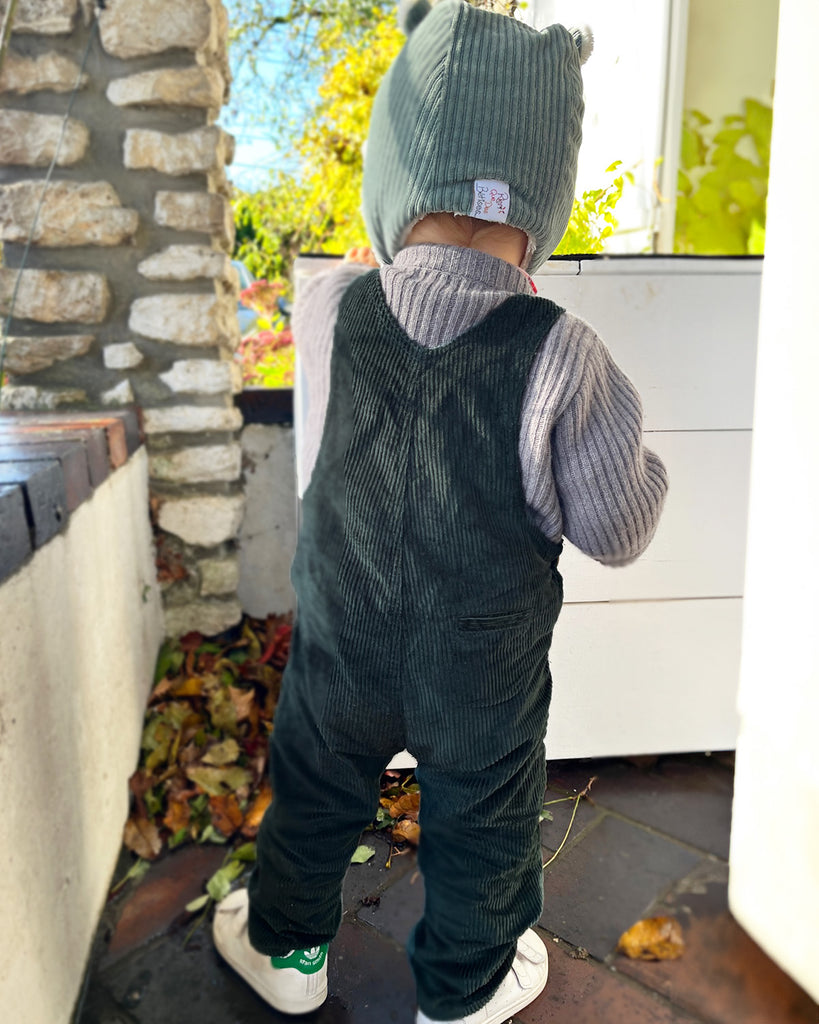 Bébé qui porte une salopette en velours vert foncé, un cardigan gris en dessous et un bonnet vert clair..